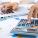 Hands using a calculator and laptop on a desk with financial charts and documents.