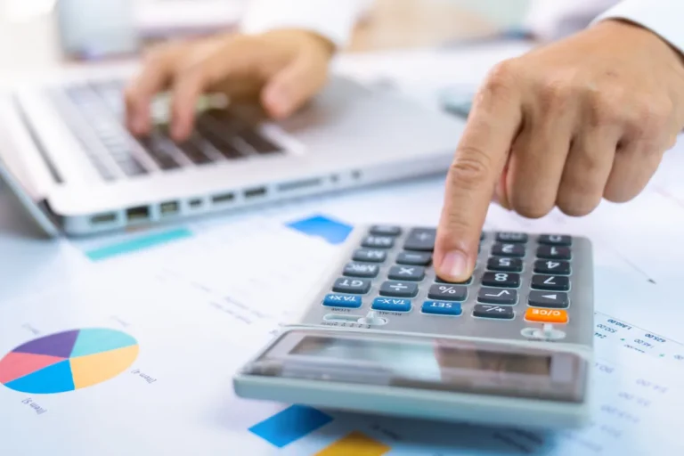 Hands using a calculator and laptop on a desk with financial charts and documents.