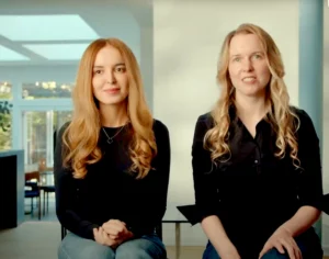 Two women with long hair sit indoors, wearing black tops, in a well-lit room with large windows in the background.