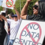 Protesters hold signs opposing data centers, one reads "No Data Center," on a sunny street with trees and buildings in the...