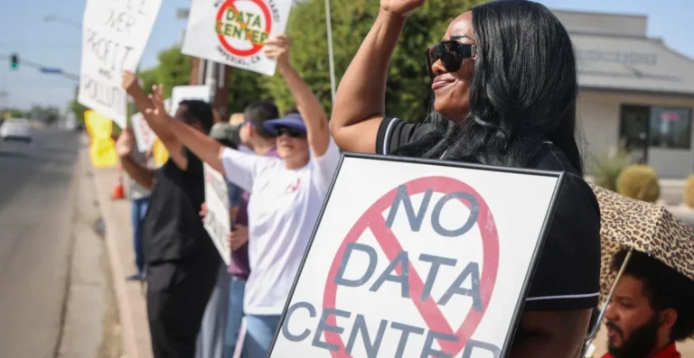 Protesters hold signs opposing data centers, one reads "No Data Center," on a sunny street with trees and buildings in the...