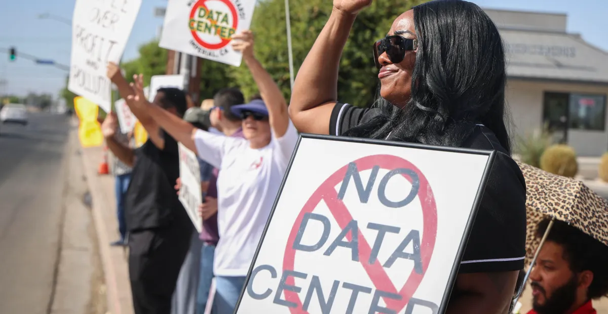 Protesters hold signs opposing data centers, one reads "No Data Center," on a sunny street with trees and buildings in the...