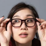 Person adjusting large black glasses, looking upwards with a neutral expression against a plain gray background.