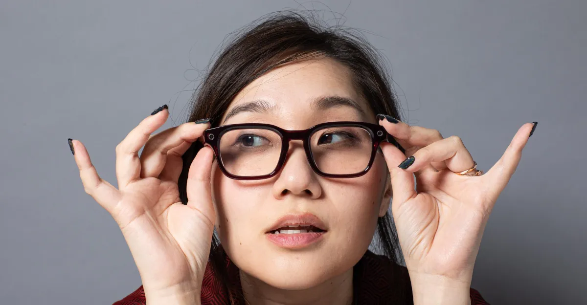 Person adjusting large black glasses, looking upwards with a neutral expression against a plain gray background.