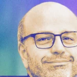 Man with glasses smiling against a gradient blue and purple background.