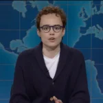 Person with curly hair and glasses speaks on a TV set with a world map backdrop.