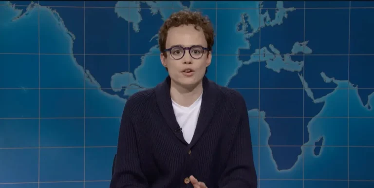 Person with curly hair and glasses speaks on a TV set with a world map backdrop.