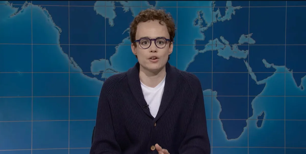 Person with curly hair and glasses speaks on a TV set with a world map backdrop.