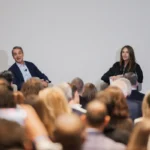 Man and woman speaking to an audience in a conference setting, with a camera recording the event.