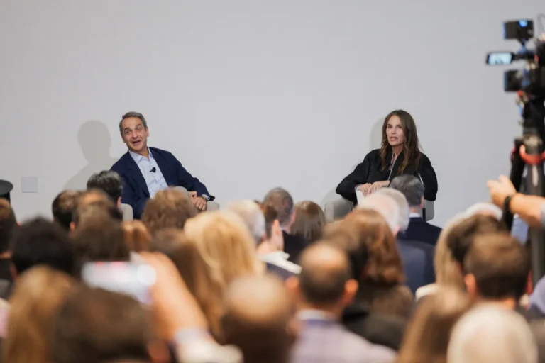 Man and woman speaking to an audience in a conference setting, with a camera recording the event.