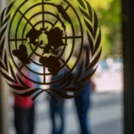 UN logo on glass door with blurred figures in the background, outdoor setting with green foliage visible.