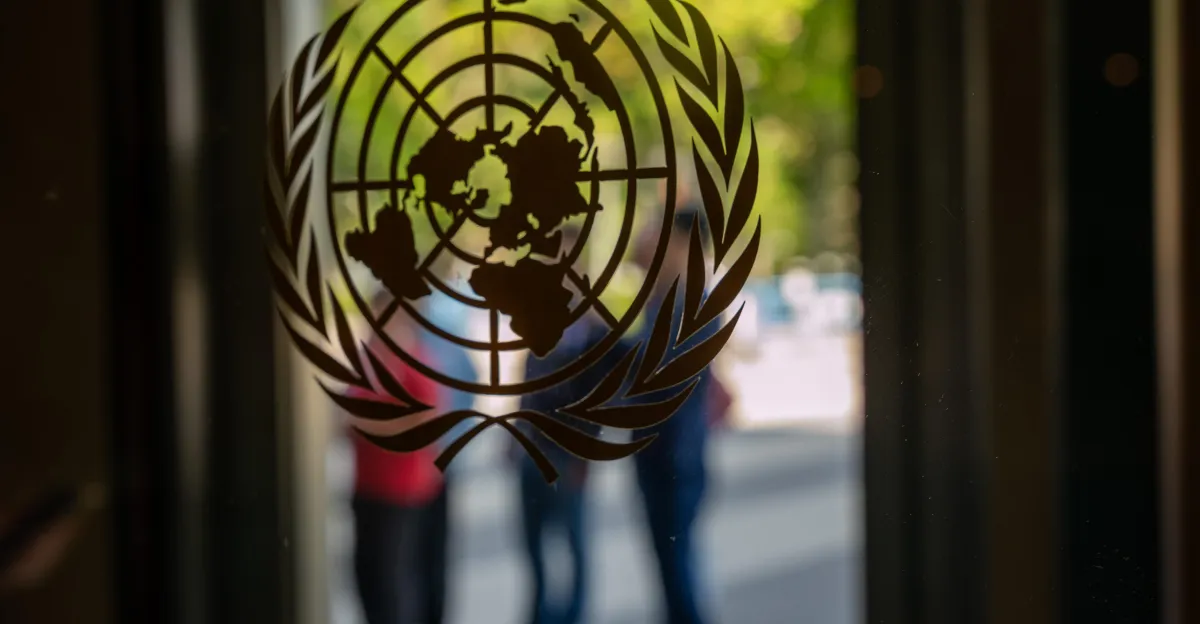 UN logo on glass door with blurred figures in the background, outdoor setting with green foliage visible.