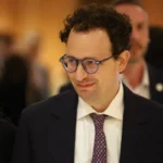 A man in a suit with glasses and curly hair smiling, standing in a crowd with a blurred background.
