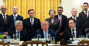 A group of executives and officials seated and standing around a table with microphones and nameplates in a formal setting.