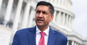 Man in a blue suit and red tie speaking outside a building with white columns in the background.
