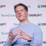 Person speaking on stage with a headset, gesturing with hands. TechCrunch Disrupt logos in the background.