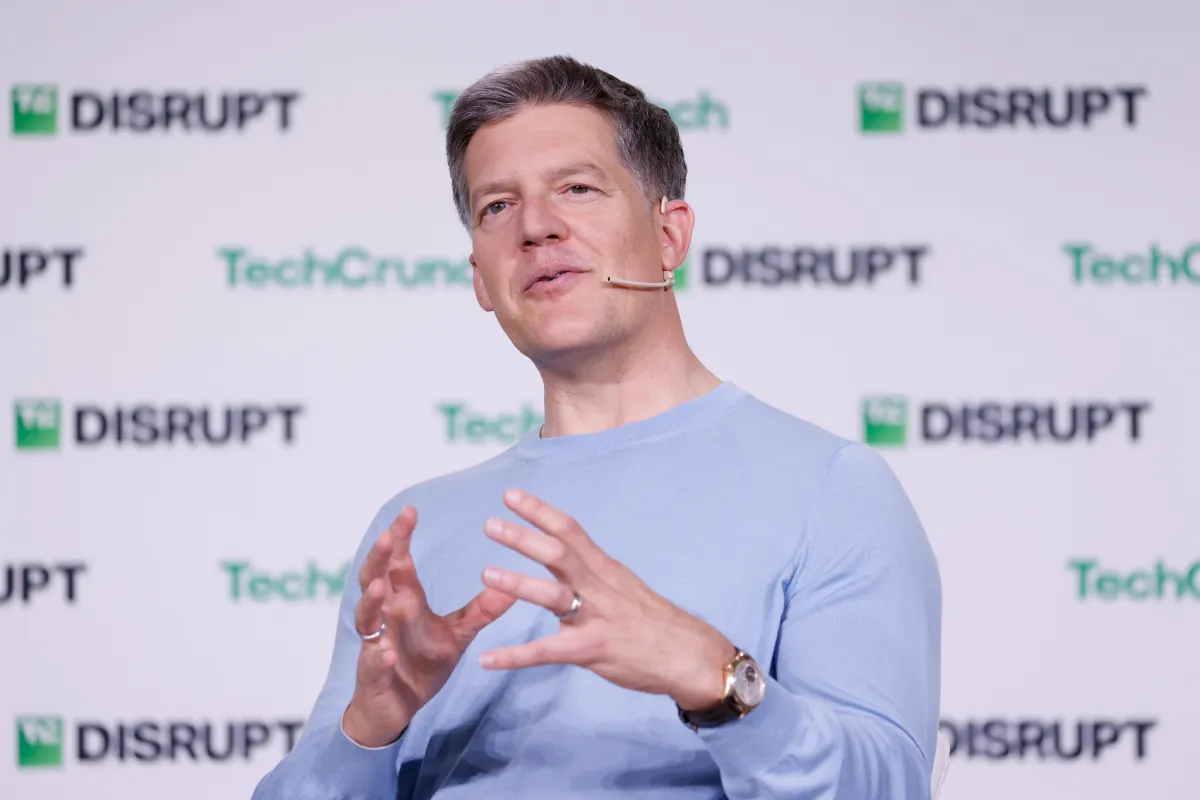 Person speaking on stage with a headset, gesturing with hands. TechCrunch Disrupt logos in the background.