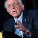 Elderly man in glasses and suit gestures while speaking at a podium, dark background.