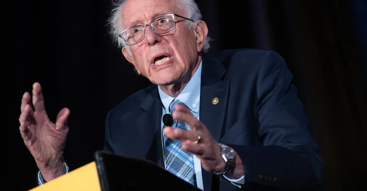 Elderly man in glasses and suit gestures while speaking at a podium, dark background.