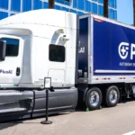 White PlusAI truck with blue NVIDIA trailer parked on a street, surrounded by palm trees and modern buildings.