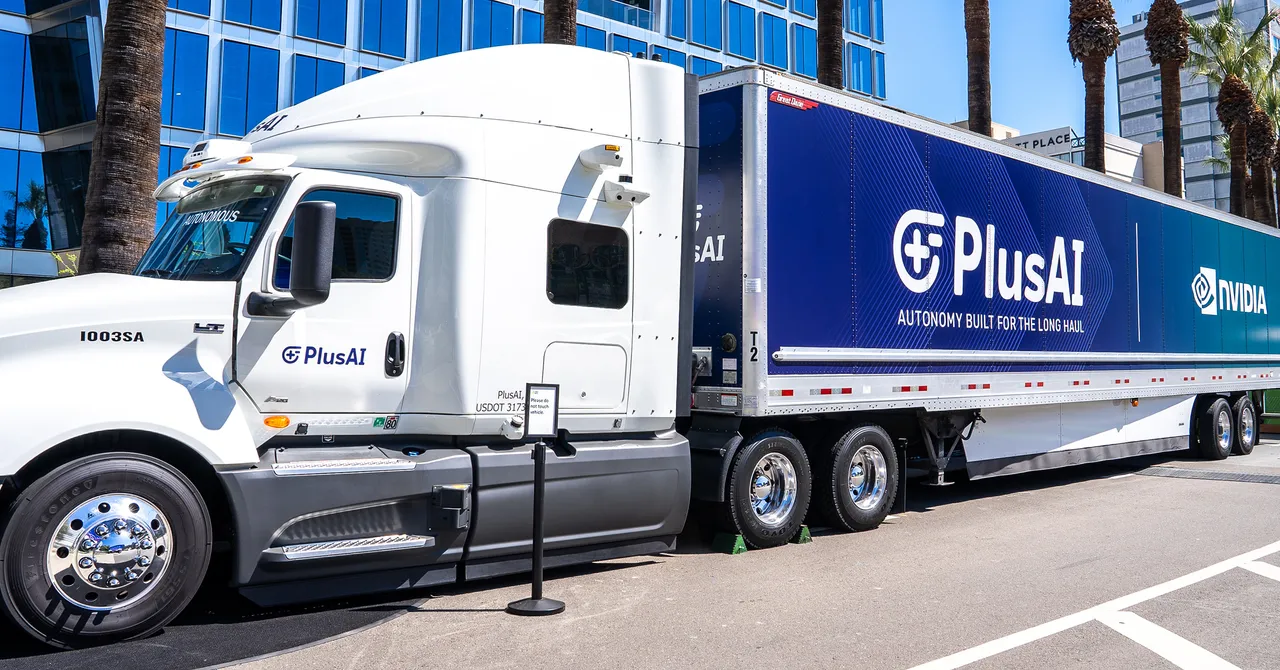 White PlusAI truck with blue NVIDIA trailer parked on a street, surrounded by palm trees and modern buildings.