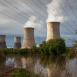 Four cooling towers emitting steam near a river, surrounded by trees, with power lines overhead under a cloudy sky.