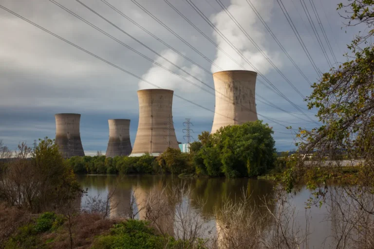 Four cooling towers emitting steam near a river, surrounded by trees, with power lines overhead under a cloudy sky.