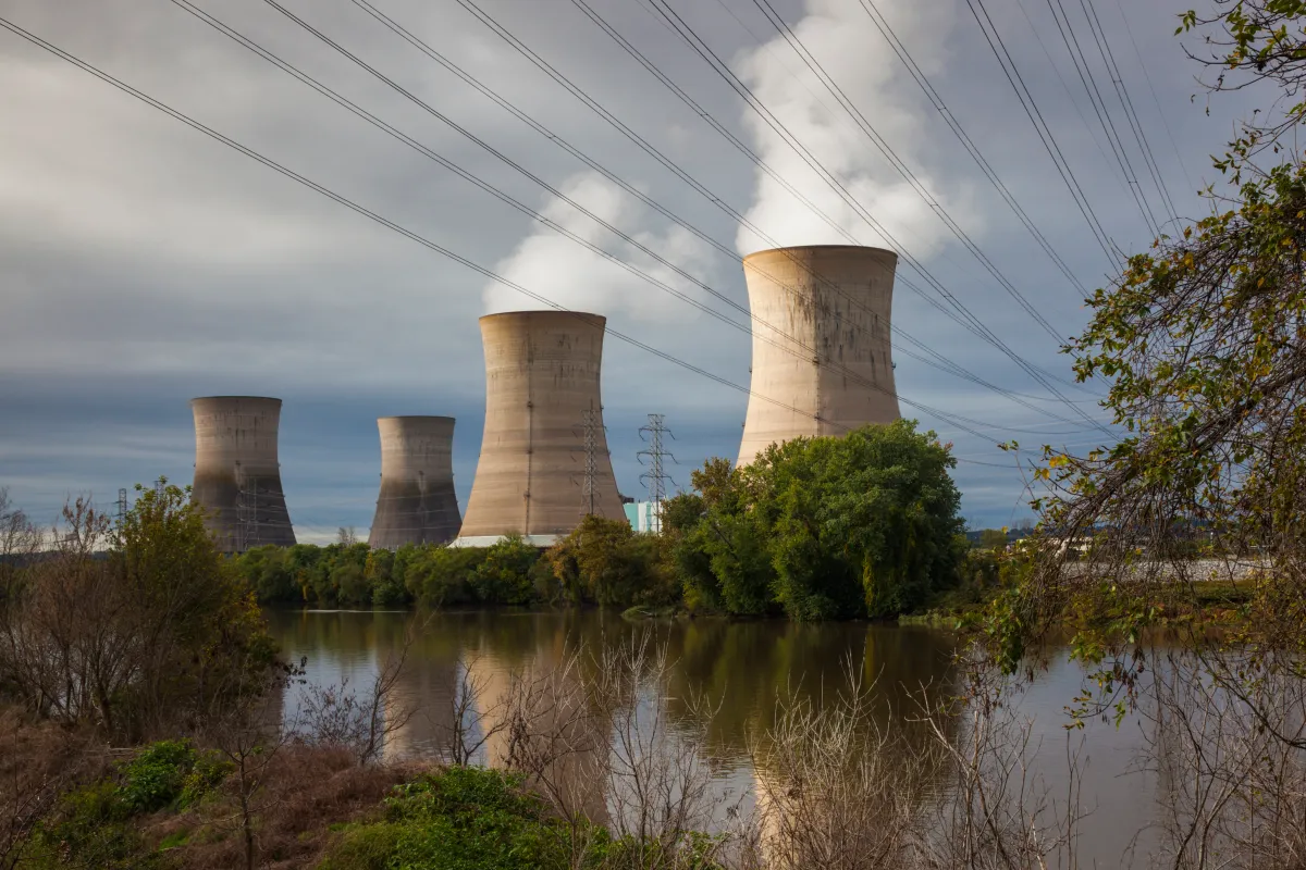 Four cooling towers emitting steam near a river, surrounded by trees, with power lines overhead under a cloudy sky.