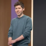 Person in a blue sweater speaking on stage with a headset, standing against a wooden and black backdrop.