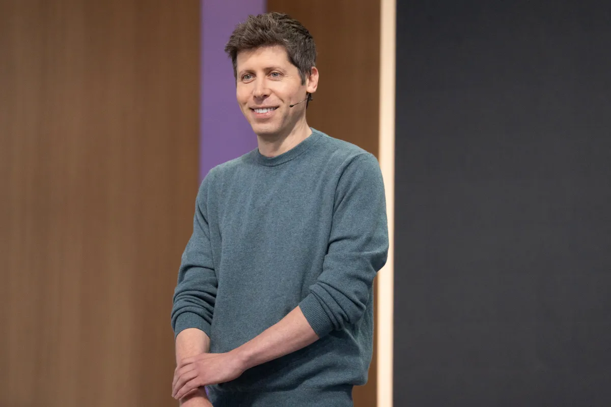 Person in a blue sweater speaking on stage with a headset, standing against a wooden and black backdrop.
