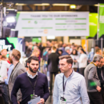 Crowd at a tech conference with banners and booths, two men in foreground wearing lanyards, visible "DISRUPT" signage.