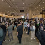 Crowded expo hall with people networking, booths displaying tech products, and overhead signs for different zones.
