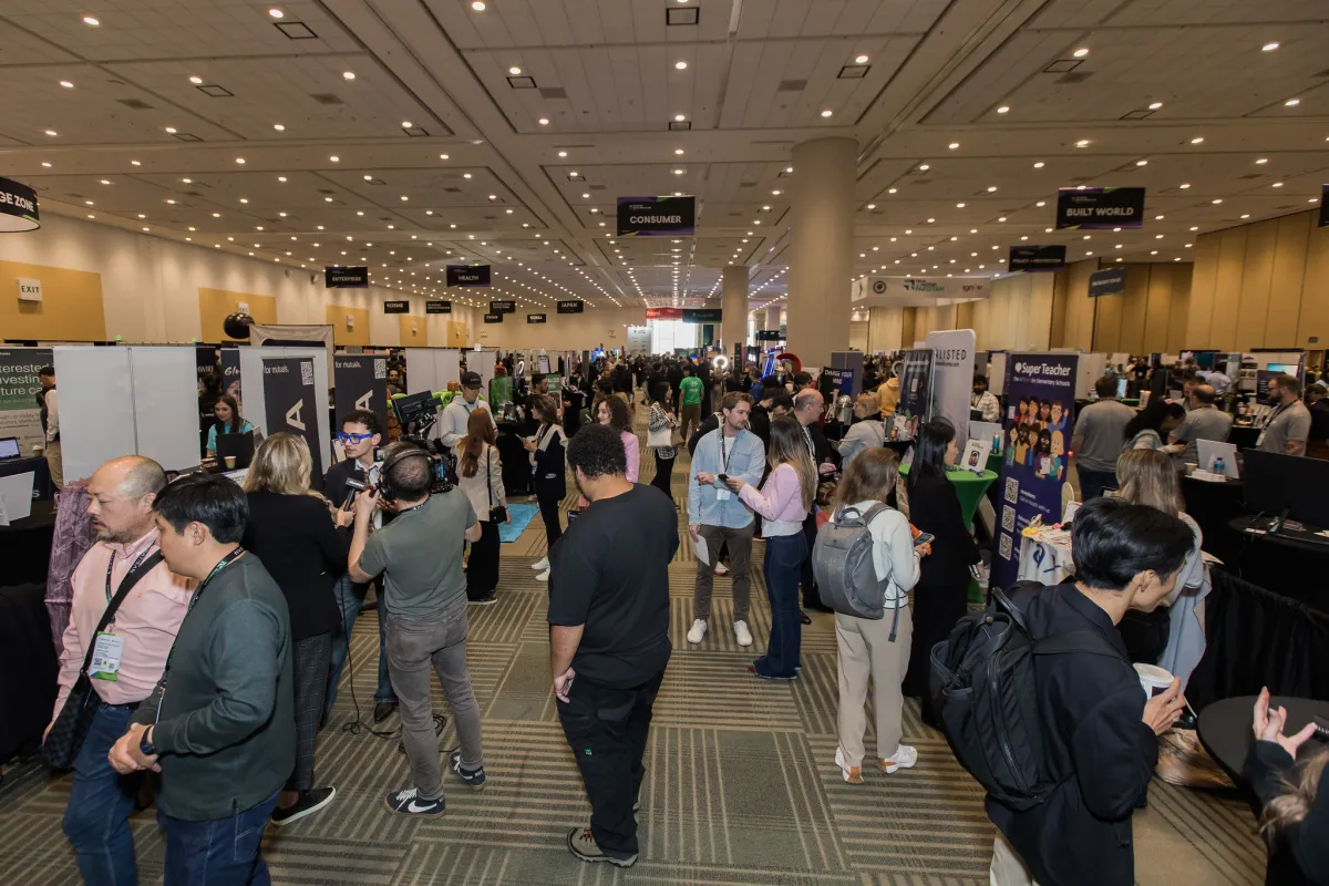 Crowded expo hall with people networking, booths displaying tech products, and overhead signs for different zones.