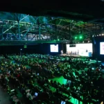 Large audience seated in a dimly lit conference hall, green lighting, stage with panelists, presentation screens visible.