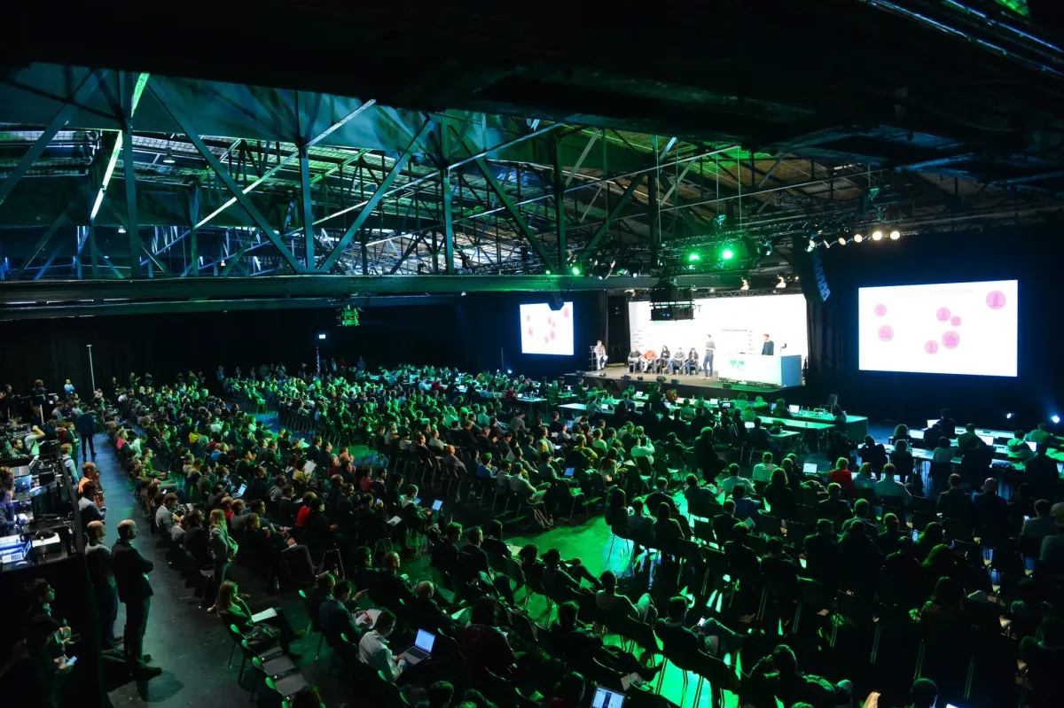 Large audience seated in a dimly lit conference hall, green lighting, stage with panelists, presentation screens visible.
