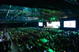 Large audience seated in a dimly lit conference hall with green lighting, watching a panel discussion on stage with screen...
