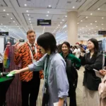 People interacting at an expo booth; a person in a tiger-striped jacket with a prize wheel; signs read "Consumer," "Built...