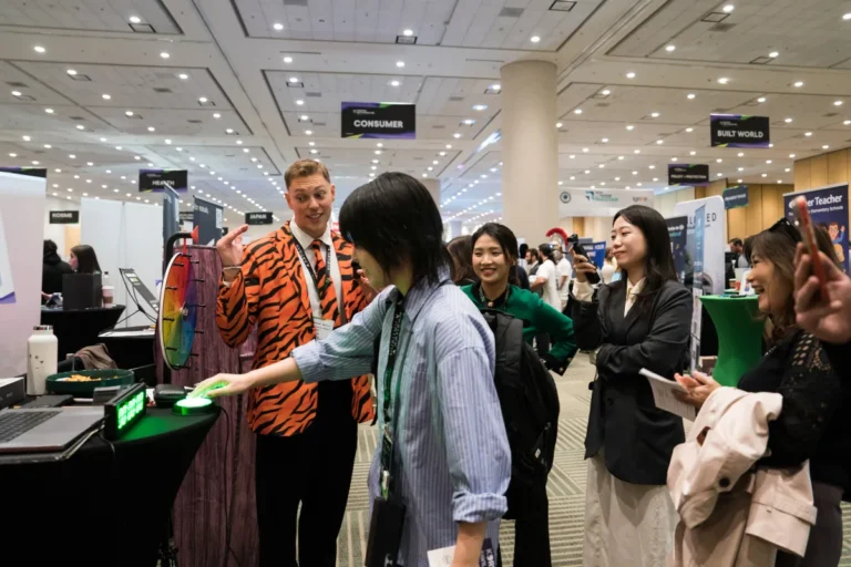 People interacting at an expo booth; a person in a tiger-striped jacket with a prize wheel; signs read "Consumer," "Built...