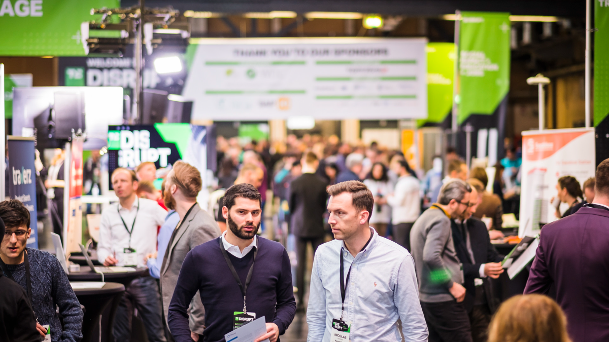 Crowd at a tech conference with banners, booths, and attendees conversing; "Disrupt" signage in background.
