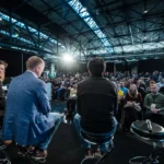 Three speakers on stage facing a large seated audience in a spacious, industrial venue with exposed beams.