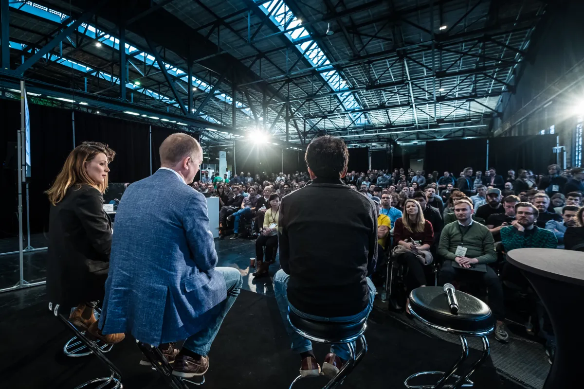 Three speakers on stage facing a large seated audience in a spacious, industrial venue with exposed beams.