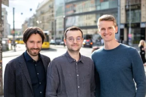 Three men standing on a busy city street with buildings and a tram in the background.