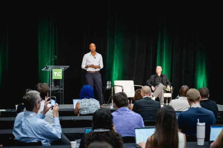 Speaker standing at podium, audience seated with laptops, green-lit backdrop, two chairs on stage, TC logo visible.