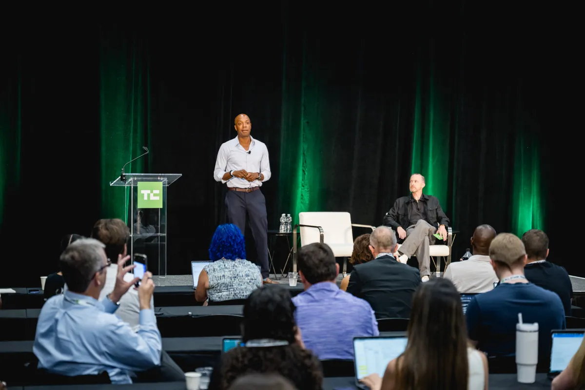 Speaker standing at podium, audience seated with laptops, green-lit backdrop, two chairs on stage, TC logo visible.