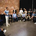 Man speaking to a seated group in a brick-walled room, attendees with lanyards, some using laptops, others taking notes.