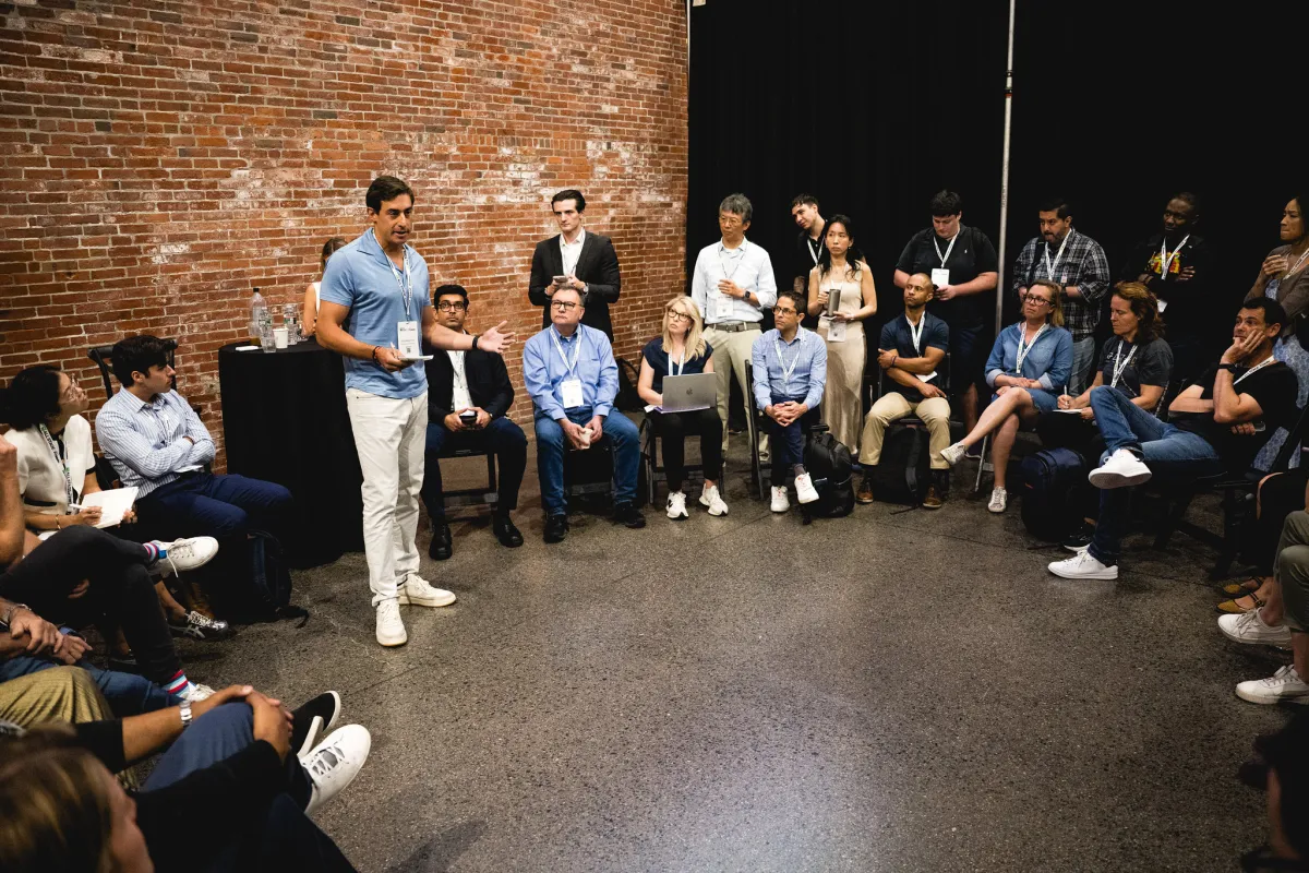 Man speaking to a seated group in a brick-walled room, attendees with lanyards, some using laptops, others taking notes.