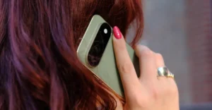 Person with red hair holding a green smartphone to their ear, wearing a silver ring and pink nail polish.