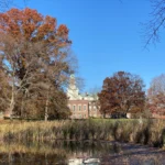 Historic building behind autumn trees and a pond under a clear blue sky.