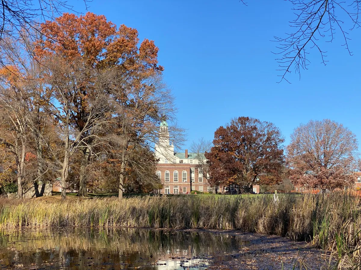 Historic building behind autumn trees and a pond under a clear blue sky.
