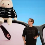 A person in a black shirt looks at a screen showing a plush toy on large eyeglasses, with a blue background.